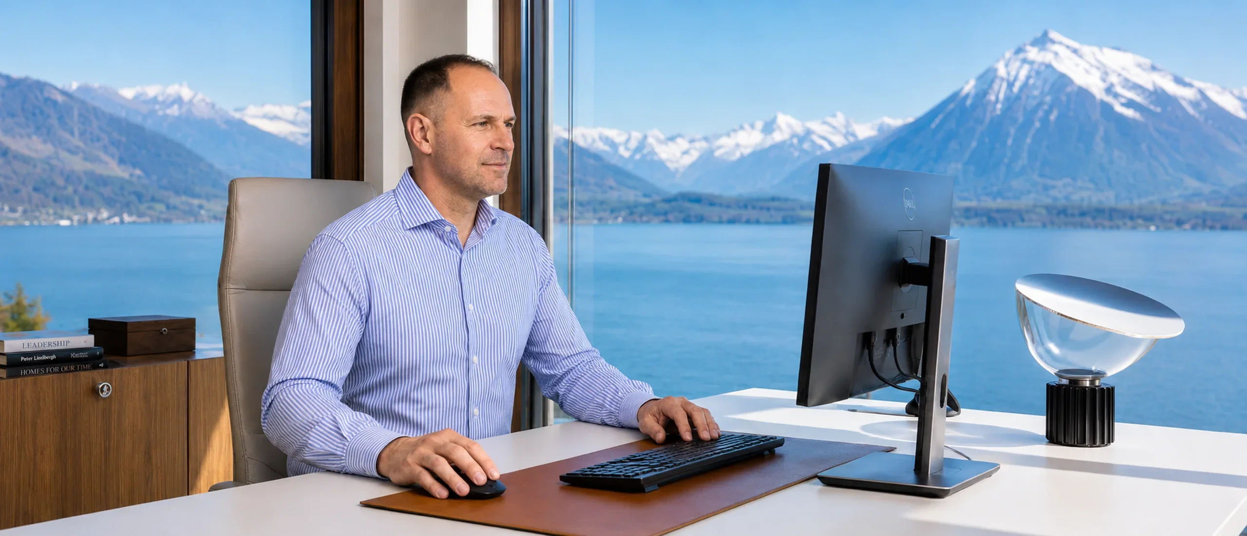 High-end, professional office scene. A man is sitting at a modern desk, seen from the side, working on a computer. Through a large window in the background, a crystal-clear, sharp view of Lake Thun and the Swiss Alps.